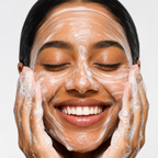 A close-up of a woman with her eyes closed, smiling broadly while washing her face with white foaming skincare cream. Her hands are also covered in the lathered cream, set against a clean, light gray background.